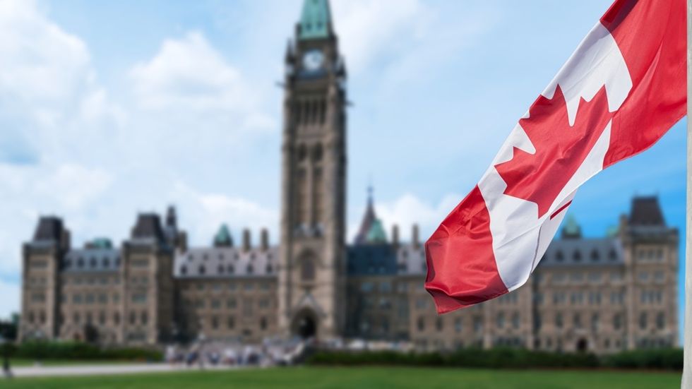 A Canadian flag waving in the foreground with the Parliament building in the background.