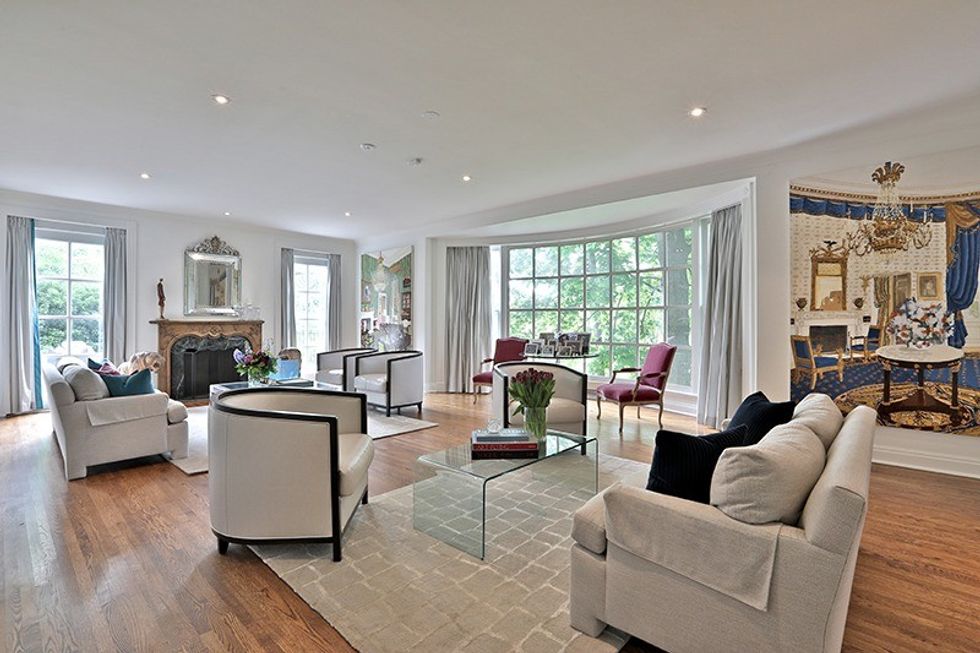 In Cohons favourite room, large bay windows allow for incredible views and light that changes with the seasons, while the chairs and throw pillows give the room a pop of colour. Family photos rest on a dainty glass table. On the right side of the window is The Blue Room painting, and on the left is The Green Room. 