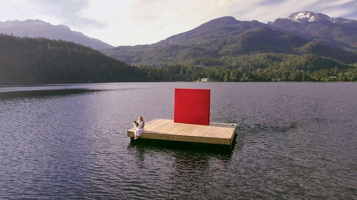 Woman and dog sitting on dock floating in lake with mountains in the background. Also on the dock is a red square, representing rennie.